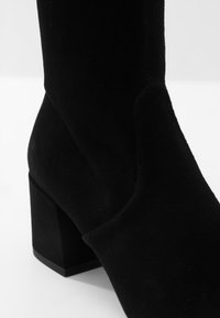 Bottine en daim noir avec talon carré, texture lisse et design sans coutures. Présente une légère découpe sur le côté pour un look moderne.