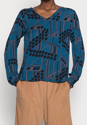 Personne portant un haut bleu à manches longues à motifs avec des points et des lignes noirs, associé à un pantalon ample beige, mains dans les poches.