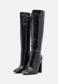 Bottes noires montantes jusqu'aux genoux en matériau texturé et brillant avec un motif peau de crocodile, un bout pointu et un talon épais. Fermeture éclair à l'arrière.