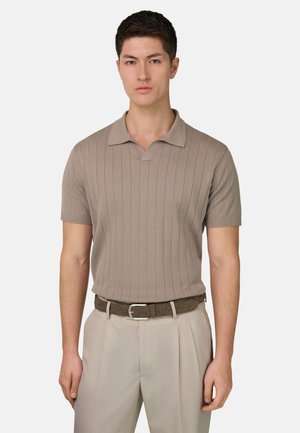 Man draagt een taupekleurig, geribd, kortermig poloshirt, ingestopt in beige geplooide broek met een bruine gevlochten riem.