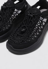 Zapatillas deportivas negras con un diseño de parte superior trenzado, cordones elásticos y una suela de goma texturizada. Presenta un logotipo en el lateral y acentos de malla.