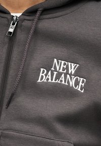 Κοντινό πλάνο ενός ατόμου που φοράει μια σκούρα γκρι μπλούζα με κουκούλα New Balance, με λευκό κεντημένο λογότυπο και μερικώς φερμουάρ μαύρου χρώματος.