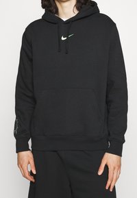 Svart hoodie med framficka och mintgrön Nike-swoosh-logotyp, buren av en person med avslappnade händer längs sidorna.