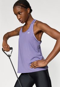 Μωβ αθλητικό τοπ με στενούς ιμάντες και λεπτομέρειες λογότυπου, συνδυασμένο με μαύρο εφαρμοστό κολάν. Ο χρήστης κρατά εξοπλισμό γυμναστικής.