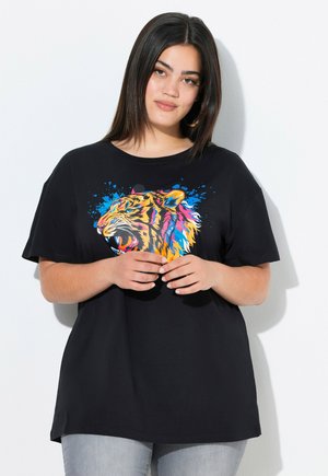 Kobieta z prostymi ciemnymi włosami, ubrana w czarny T-shirt z kolorową grafiką ryczącego tygrysa, stojąca na białym tle z złożonymi dłońmi.