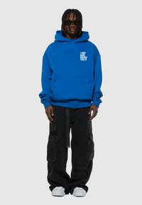Blå huvtröja med en framficka i kängurustil och "LOST YOUTH" tryckt i vitt. Matchas med svarta cargobyxor och vita sneakers.