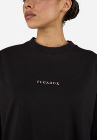 Black sweatshirt featuring a crew neck and a subtle pink "PEGADOR" logo on the chest. Soft fabric with a relaxed fit and minimal design.