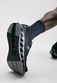 Schwarzer Sportschuh mit einem strukturierten Obermaterial und einer grauen, gepolsterten Sohle, die Luftpolster sowie ein markantes Profilmuster aufweist.