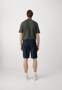 Camicia a maniche corte di un verde oliva testurizzato abbinata a pantaloni corti blu navy, rifinita con orli arrotolati e sneakers bianche.