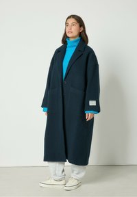 Námornícky vlnový oversize kabát s dvoma prednými vreckami, s jedným gombíkom, v kombinácii s jasne modrým rolákom a šedými teplákmi.