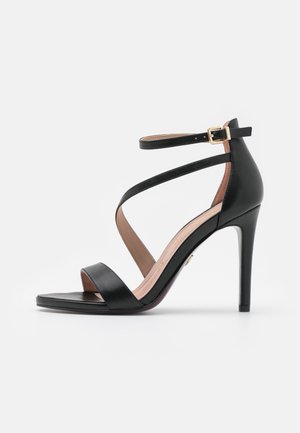 Sandale noire à talons hauts avec un bout carré, deux larges sangles sur le pied et une sangle ajustable à la cheville avec une boucle en or.