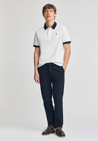 Hvid polo skjorte med marineblå krave og ærmekanter, parret med mørkeblå bukser og brune sko med orange snørebånd. Slim fit design.
