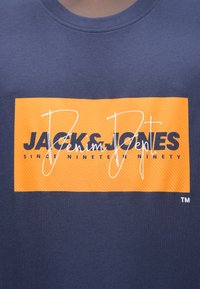 Μπλε ναυτικού χρώματος t-shirt με πορτοκαλί ορθογώνιο γραφικό με υφασμένες ρίγες, που εμφανίζει "JACK & JONES" και "Denim Dept" σε αντίθετες γραμματοσειρές.