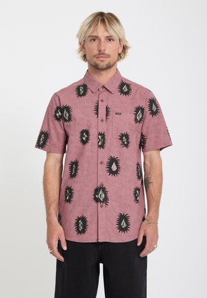 Homme blond portant une chemise à manches courtes rose poussiéreux à boutons avec des motifs géométriques épineux noirs et gris, et un pantalon noir.