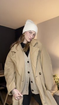 Bež oversize jakna od gornjeg dijela tijela s visokim ovratnikom i dvostrukim redom gumba, nošena preko teksturiranog bež kardigana i crne majice, u kombinaciji s bijelom pletenom kapom.