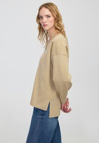 Beżowa, luźna bluzka z dekoltem w serek i szerokimi rękawami, z bocznymi rozcięciami i teksturowanym splotem. Noszona z niebieskimi dżinsami.
