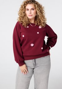 Vrouw met krullend blond haar, gekleed in een bordeauxrode trui versierd met roze bloemenapplicaties en lichtgrijze jeans, staand tegen een witte achtergrond.