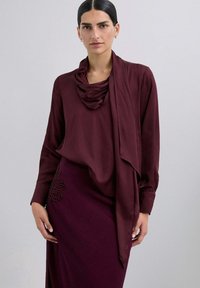 Blusa burdeos con escote drapeado y mangas largas, combinada con una falda a juego que presenta detalles de cuentas florales. Tela suave y texturizada.