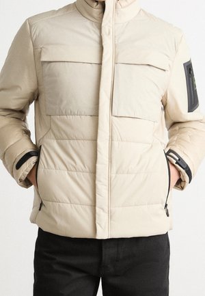 Beige quilted jacket with zippered front, flap chest pockets, zippered side pockets, and adjustable wrist straps worn by person with hands in pockets.