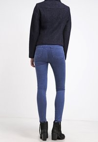 Osoba ubrana w ciemny wełniany żakiet, wąskie niebieskie dżinsy i czarne botki za kostkę, stojąca tyłem na jednolitym białym tle.