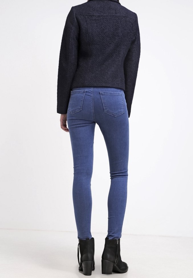 Osoba ubrana w ciemny wełniany żakiet, wąskie niebieskie dżinsy i czarne botki za kostkę, stojąca tyłem na jednolitym białym tle.