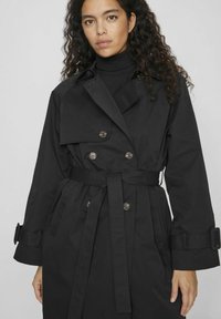 Svart trenchcoat med dubbelknäppt design, bälte och knappdetaljer. Har en medellång snitt och skräddarsydd passform. Slät tygtextur.