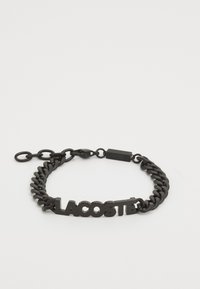 Crna metalna lancana narukvica s odvažnim, uzdignutim logotipom "LACOSTE" i sigurnom kopčom. Dizajn ima glatku, sjajnu teksturu.