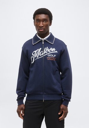 Homme portant une veste zippée bleu marine avec les textes « Malbon Golf » et « Always On The Hunt Los Angeles, CA » sur le devant, debout devant un fond blanc.
