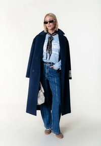 Dolgi mornarsko modri plašč čez svetlo modro majico s potiskom belih črk, kombiniran z razširjenimi hlačami z modro-belim črtastim vzorcem in rjavimi obutvami brez vezalk. Vključen je tudi črni nakit.