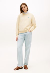 Εκρού φούτερ με μπλε λογότυπο, σε συνδυασμό με ανοιχτό μπλε φαρδιά τζιν παντελόνια και καφέ σανδάλια. Απλός σχεδιασμός, μαλακό υλικό, άνετη εφαρμογή.