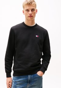 Black sweatshirt with a ribbed crew neck, featuring a small embroidered logo on the chest. Paired with blue denim jeans.