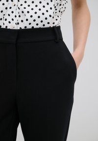 Black tailored trousers with a textured fabric, featuring a belt looped waistband and side pockets. Paired with a white polka dot shirt.