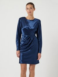 Nő, aki fényes tengerészkék hosszú ujjú, átlapolt ruhát visel, derékán redőzött részlettel, egyszerű világos háttér előtt áll.