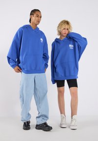 Felpe oversize blu con tasche frontali e dettagli del logo, abbinate a jeans blu chiaro e pantaloni da bici neri, completate da sneakers bianche.