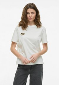 Camiseta de algodón blanca con cuello redondo, mangas cortas y un gráfico de labios con estampado de leopardo en el lado izquierdo del pecho. Lleva pantalones gris oscuro.