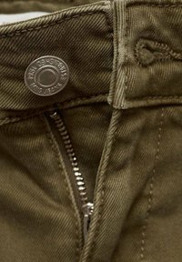 Jean kaki avec fermeture éclair partiellement ouverte sous un bouton métallique marqué "King Jeans Established 1984", avec des coutures visibles et la texture du tissu.