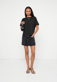 T-shirt preta de mangas curtas com um logotipo, combinada com uma saia em ganga preta com bolsos frontais. A modelo usa sandálias com tiras transparentes.