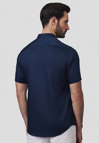 Granatowa koszula z krótkim rękawem o gładkiej fakturze, z kołnierzykiem zapinanym na guziki i projektowanym wycięciem na plecach. Dopasowany krój z zaokrąglonym dołem.
