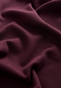 Hlboká bordová látka s hladkým, textúrovaným povrchom, ktorá má jemné záhyby a závesy, čím zdôrazňuje svoju mäkkosť a pružnosť.