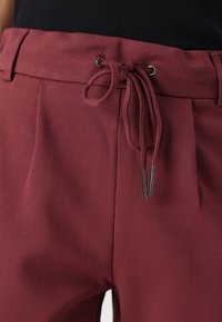 Bordeauxfärgade byxor med dragsko i midjan och metalldetaljer, tillverkade av ett mjukt tyg med skräddarsydd passform och veckdetaljer.