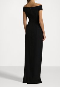 Žena oblečená v dlouhých černých šatech s odkrytými rameny, zobrazená zezadu na jednobarevném světlém pozadí.