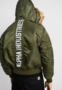 Maslinasto zeleni zimski kaput s kapuljačom, koji ima lažni krzneni obrub, rebraste rukave i veliki bijeli logotip "ALPHA INDUSTRIES" na leđima.