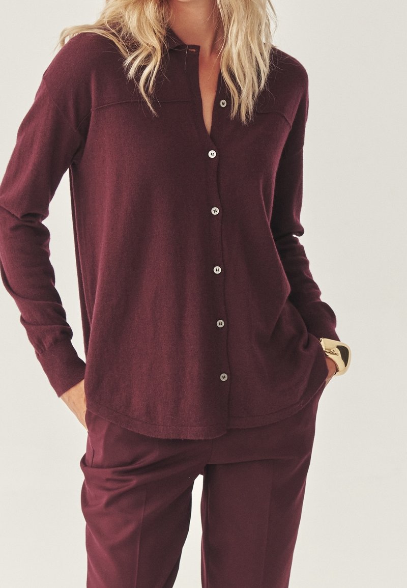 Femme portant un haut bordeaux à manches longues avec boutons et un pantalon assorti, une main dans la poche et un bracelet en or au poignet.