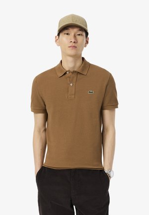 Polo shirt marrone con colletto, maniche corte e logo ricamato verde. Abbinato a pantaloni in corduroy scuri e un cappellino beige.