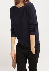 Pull côtelé marine avec un large décolleté et des manches longues, associé à un pantalon noir et une pochette de couleur claire à la main.