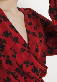 Blouse rouge à motifs floraux noirs, design à décolleté en V profond, tissu drapé et manches longues. Texture douce avec une coupe décontractée.