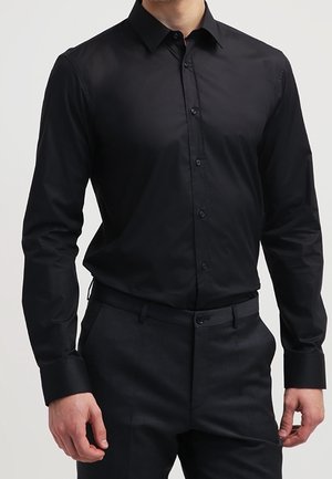 Man in een passend zwart overhemd met knopen, ingestopt in zwarte pantalon, met lange mouwen en een standaard kraag.