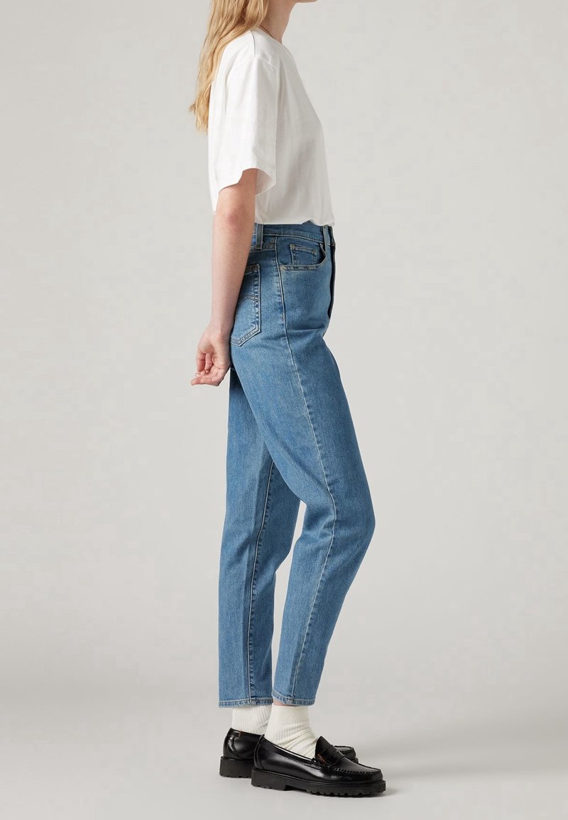 Femme debout de profil, portant une chemise blanche à manches courtes, un jean bleu taille haute, des chaussettes blanches et des mocassins noirs sur fond gris.
