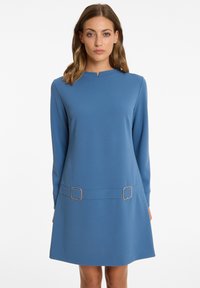Blaues langärmliges Kleid mit rundem Ausschnitt, ausgestattet mit zwei goldenen quadratischen Akzenten in der Taillenregion und einer lockeren Silhouette. Weiche Textur.