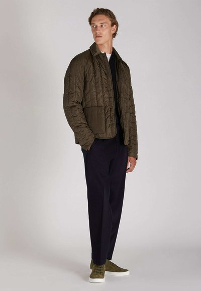 Veste matelassée marron avec col, fermeture à boutons, et deux poches avant, portée sur un haut sombre et un pantalon sombre ajusté avec des baskets vertes.